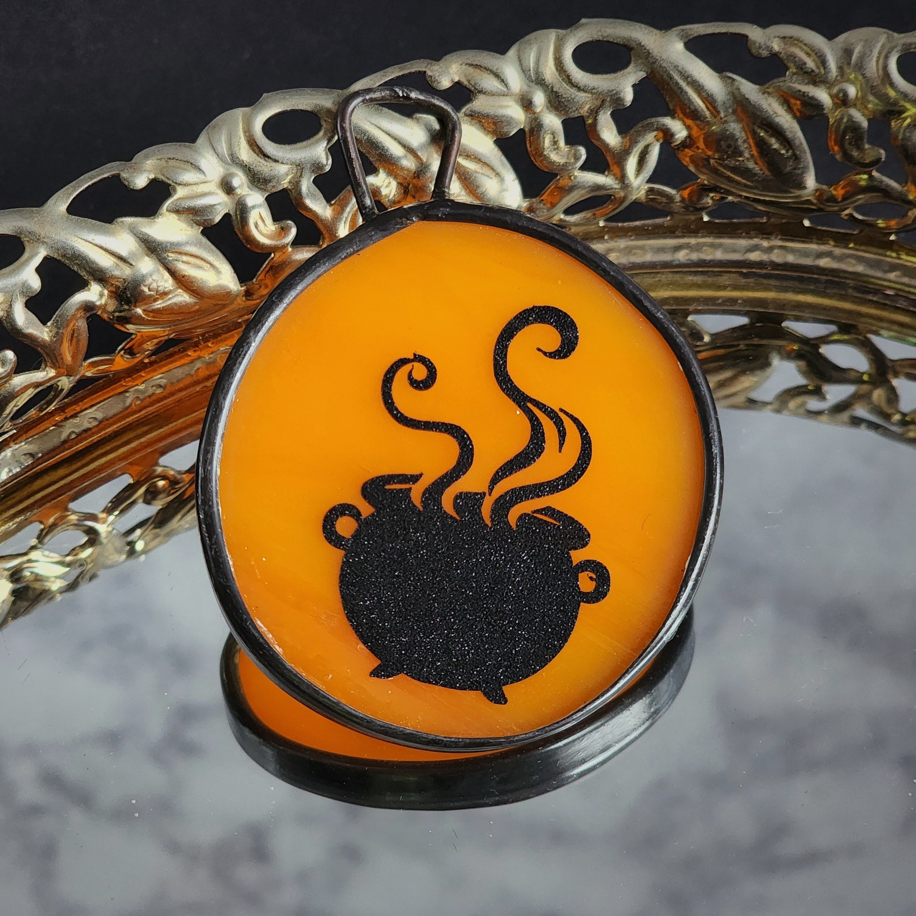 Black silhouette of a cauldron with steam on an orange stained glass, displayed on a decorative background