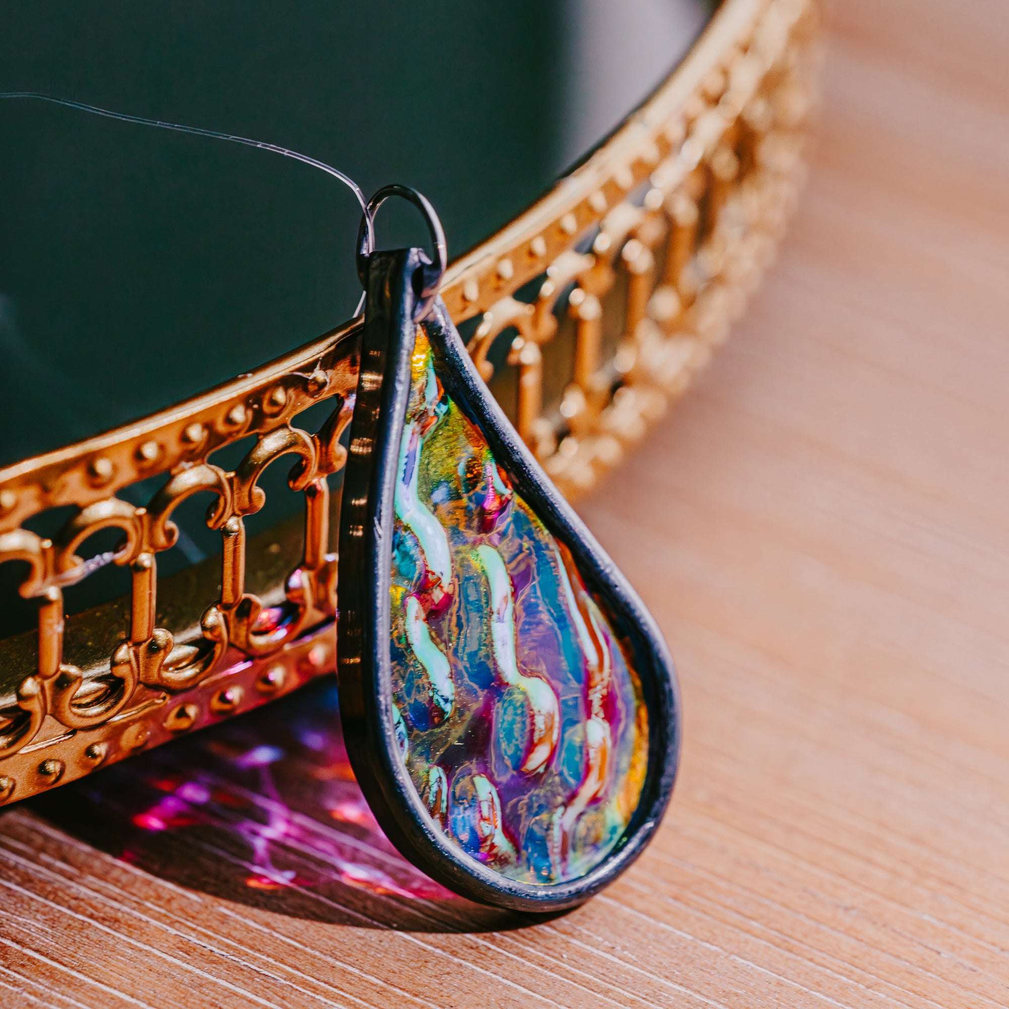 Colorful teardrop-shaped car charm on a wooden surface with a decorative metal object in the background.