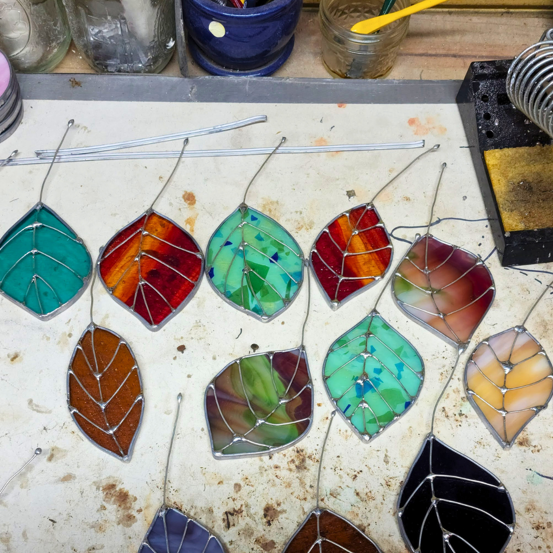 A collection of colorful stained glass leaf pieces laid out on a workbench next to a soldering iron and other crafting supplies.