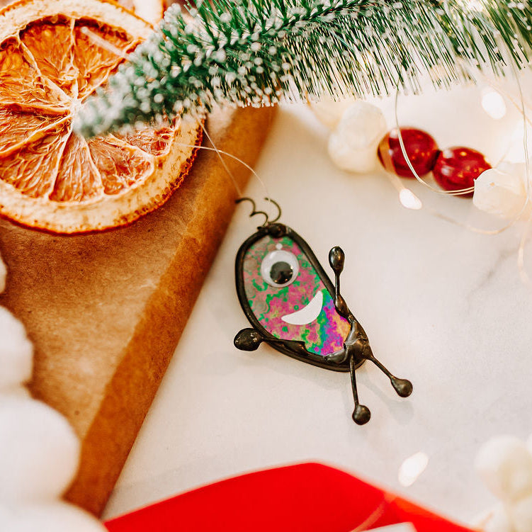 A scrappy creature made if iridescent stained glass, displayed on a kraft paper box with dried orange slices, a miniature pine branch, white pom-pom garland, red ribbon, and warm string lights.