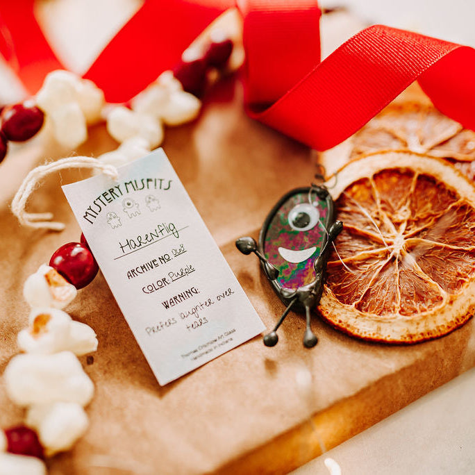 A wrapped gift box tied with a red ribbon, decorated with dried orange slices, a small decorative scrappy glass character, and a white adoption card