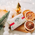 A wrapped gift box labeled 'Mystery Mum' sits on a white background surrounded by holiday props, including dried orange slices, a small green tree, white fluffy garland, and warm string lights.