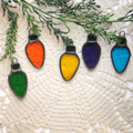 Five Old-Fashion Christmas lightbulb shaped ornaments in 5 colors (green, orange, yellow, purple, and blue) arranged on a cream lace doily with green pine branches.