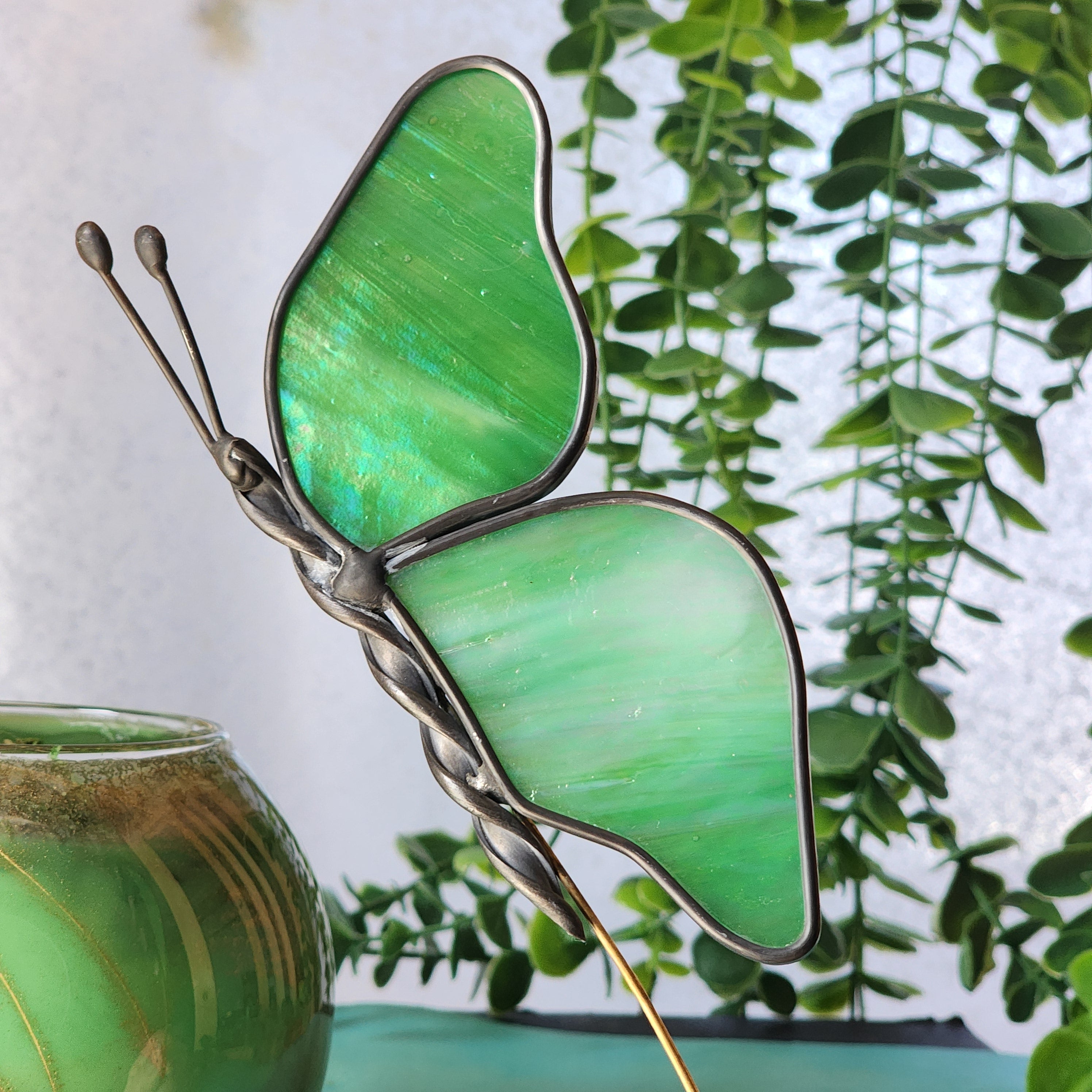 Green butterfly-shaped plant stake with an iridescent body against a blurred green leafy background
