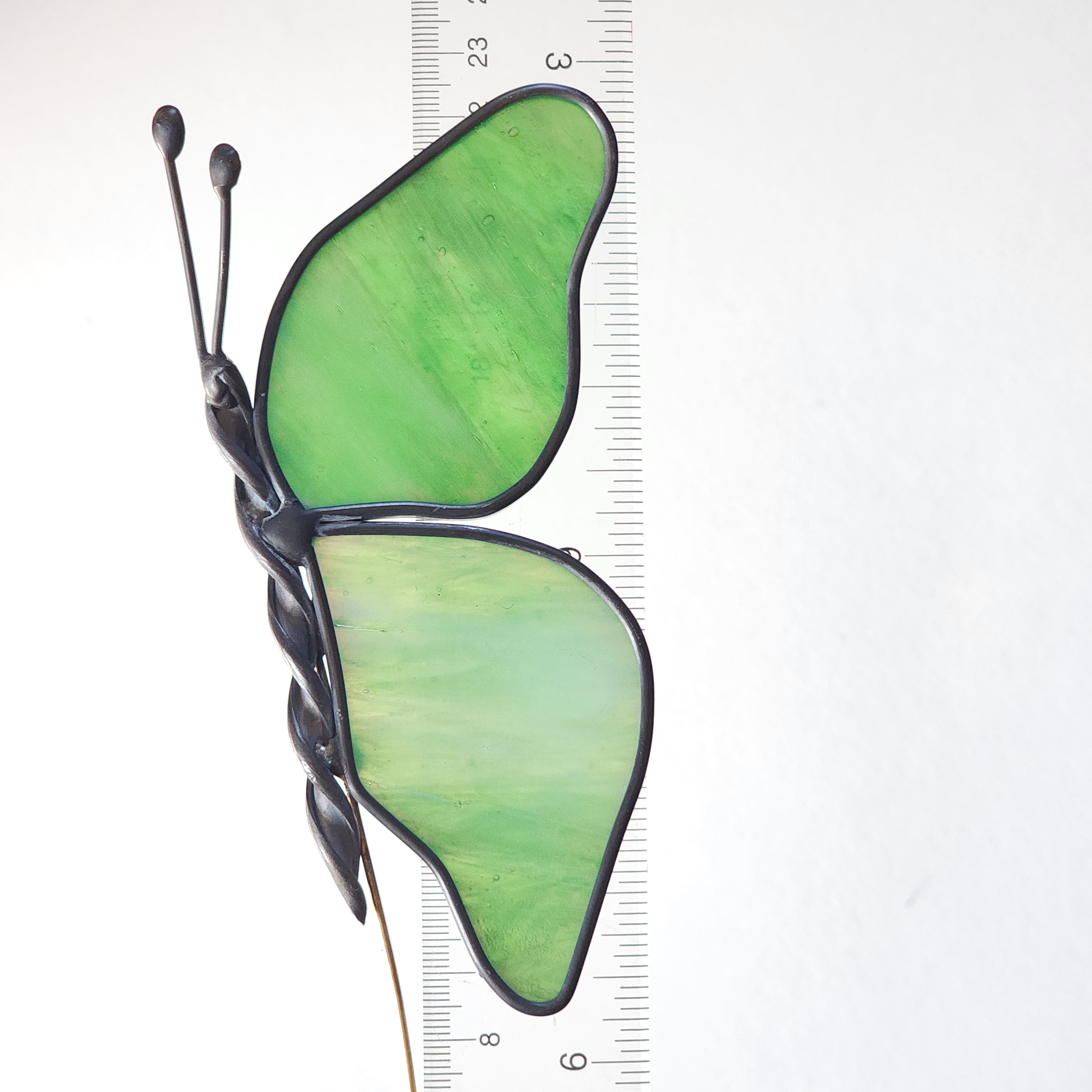 Butterfly-shaped plant stake ornament with green wings and black body on a white background with a ruler for scale.