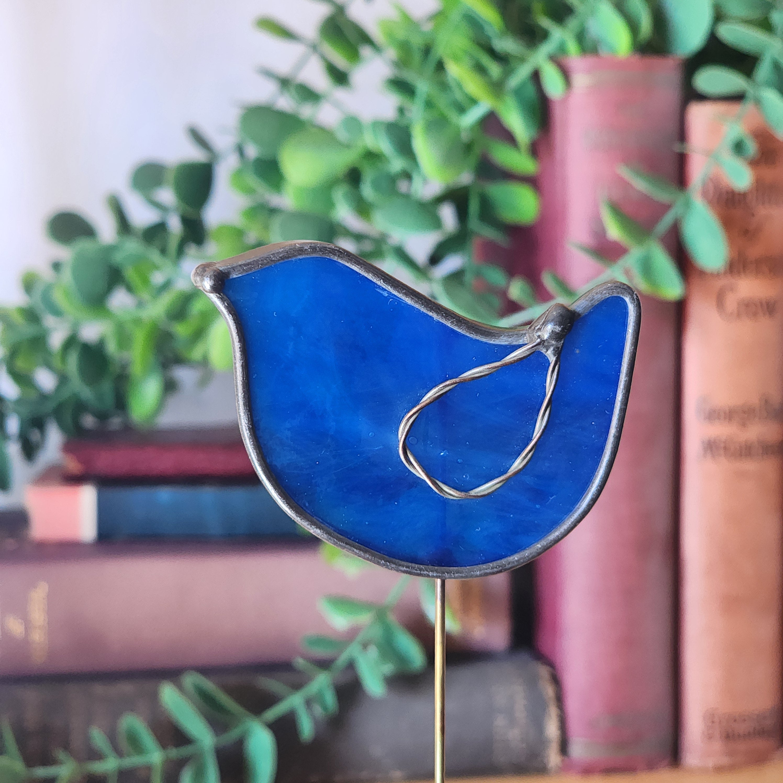 Blue bird stained glass plant stake with wire twist wings against a blurred bookshelf and greenery background
