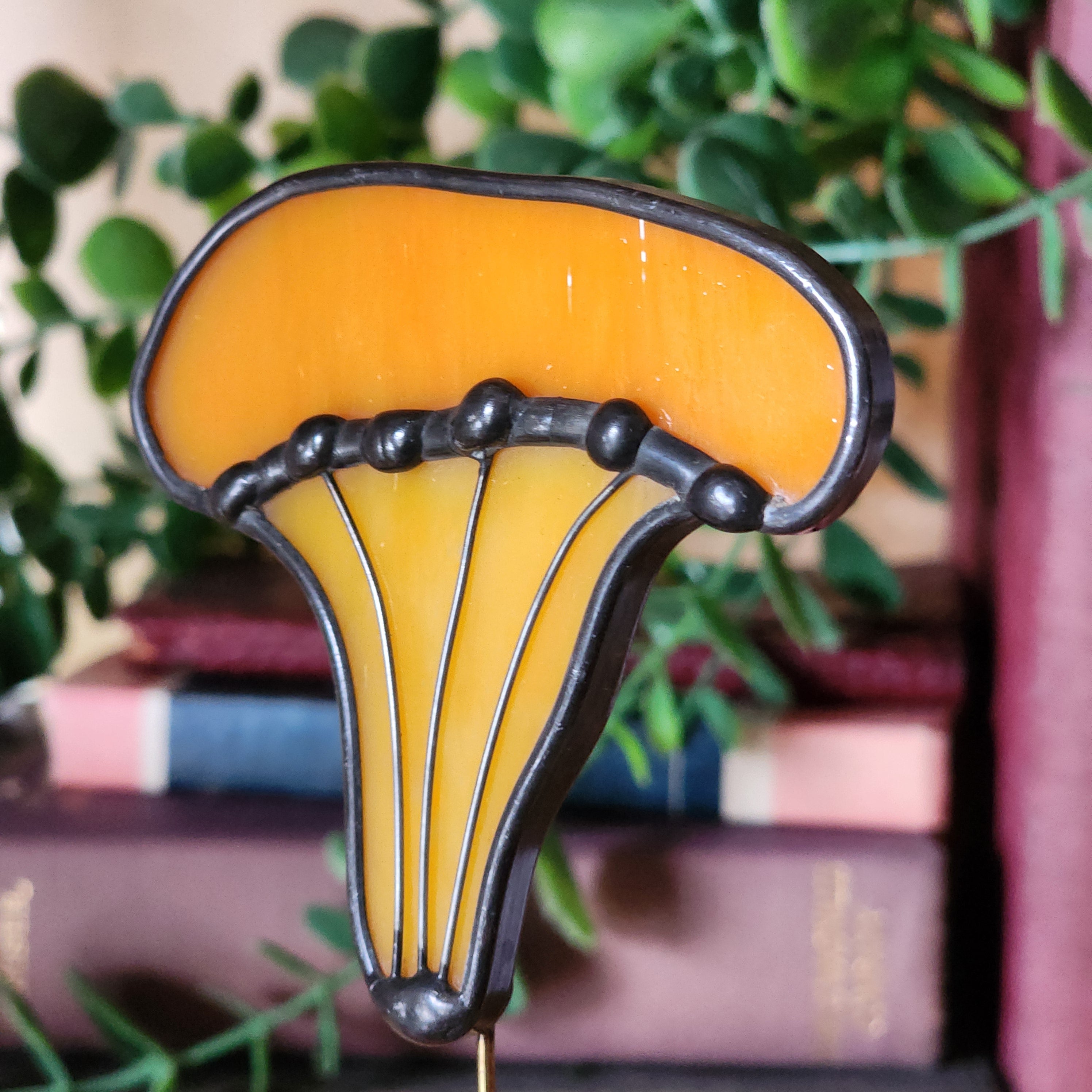 Decorative stained glass chanterelle mushroom plant stake, set against a blurred background of books and greenery.