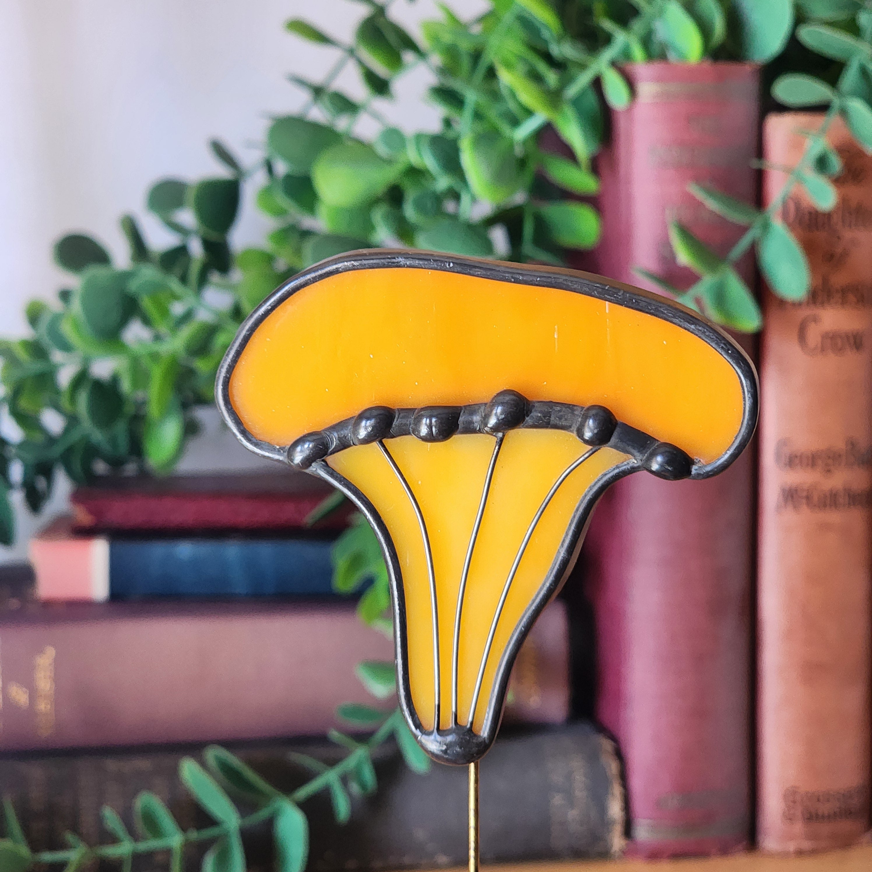 Decorative chanterelle plant stake in yellow orange stained glass, on a background of books and greenery