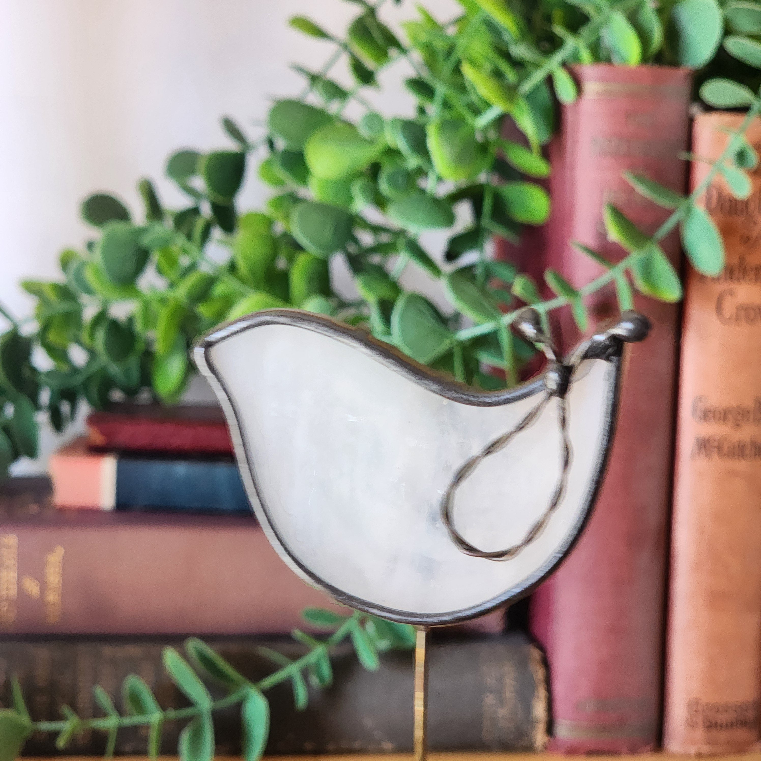 Decorative white stained glass dove plant stake on a background of stack of books with greenery