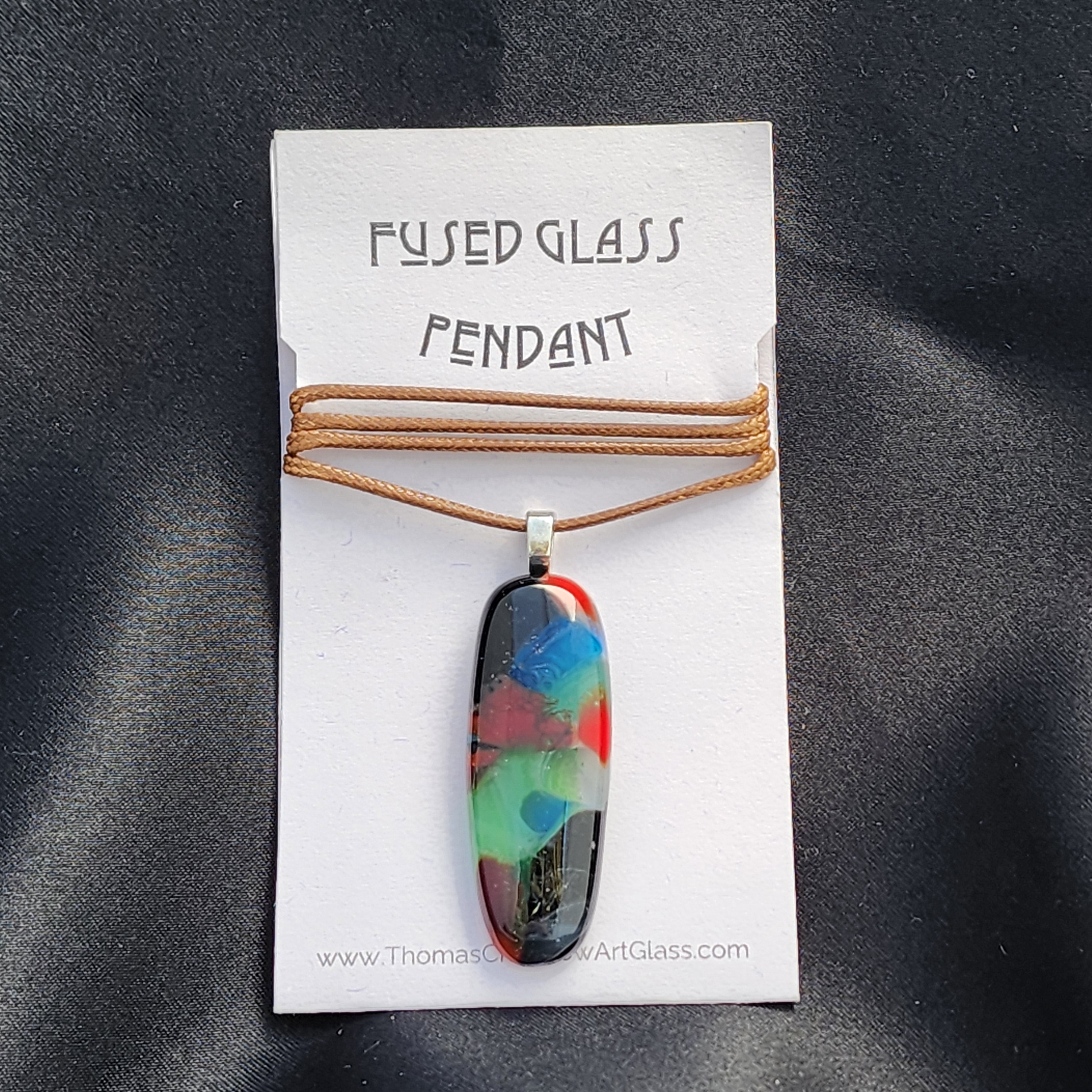 Fused glass pendant on a card with 'Thomas Glass' branding against a dark fabric background