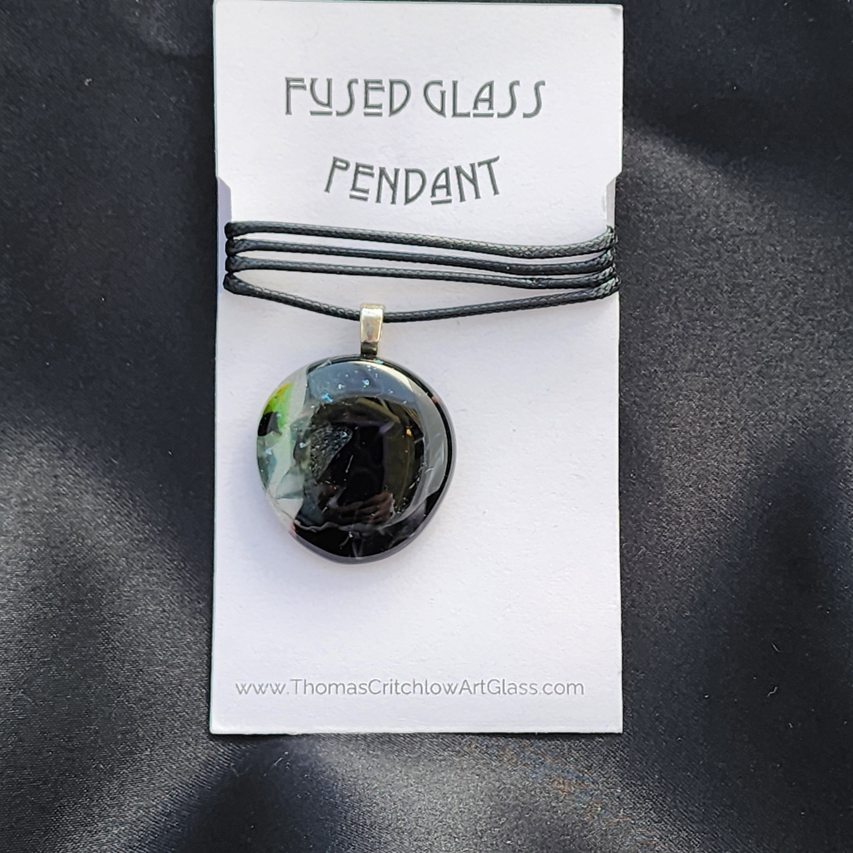 Fused glass pendant on a black cord with a label on a dark gray background