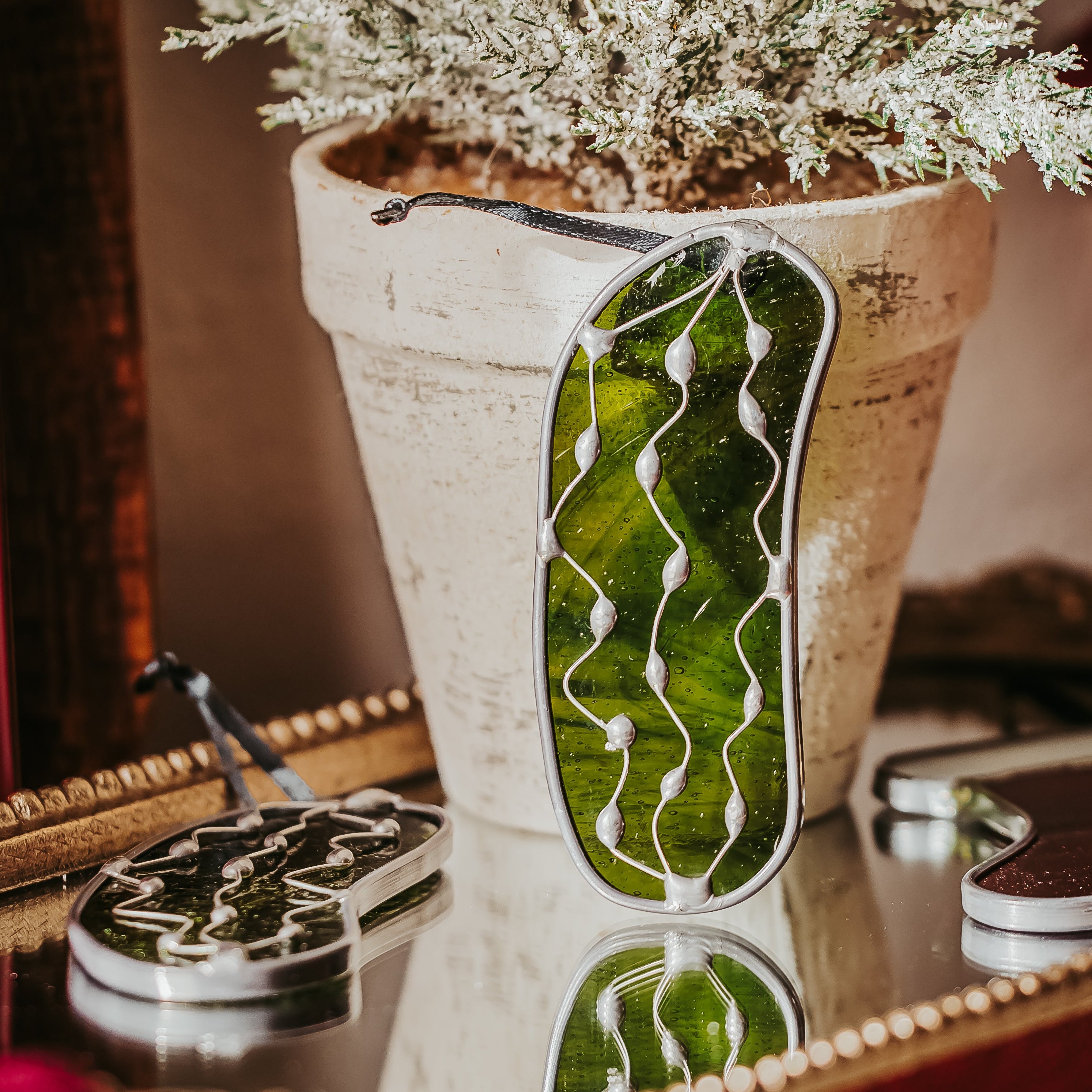 Decorative green Christmas Pickle on a mirror with a plant in the background.