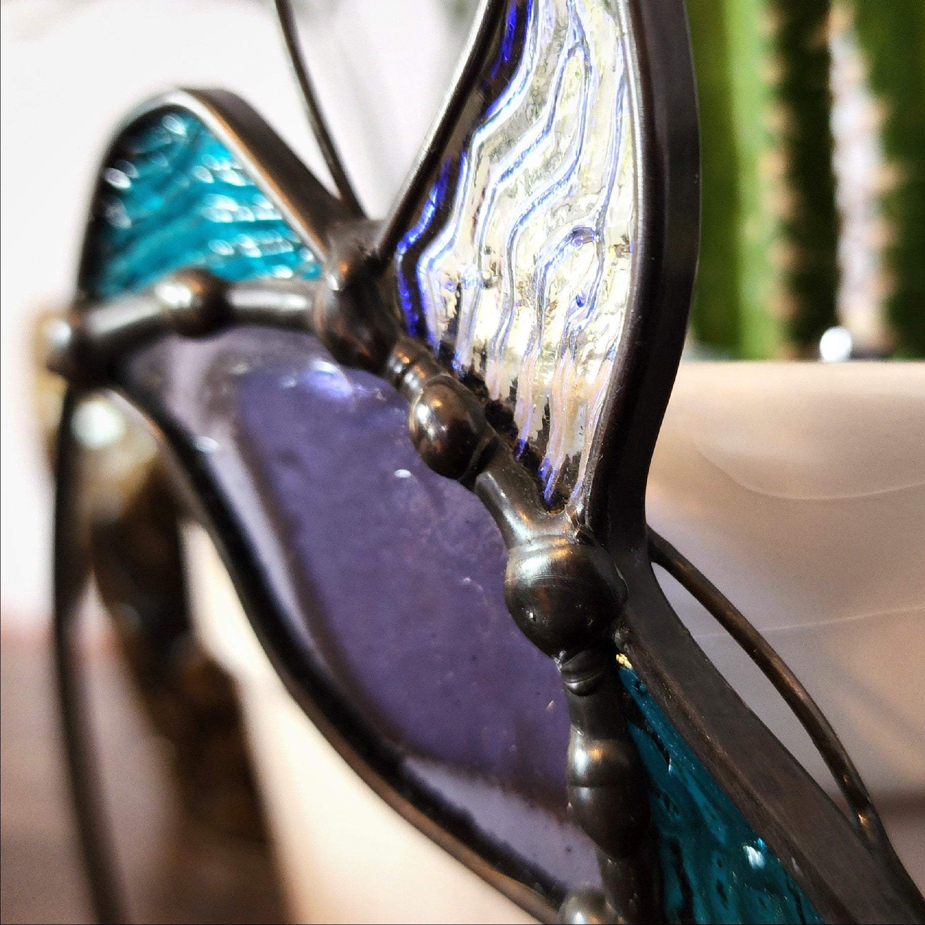A close-up of a stained glass hummingbird  ornament with purple, teal blue, and silver glass panels separated by dark metal solder.