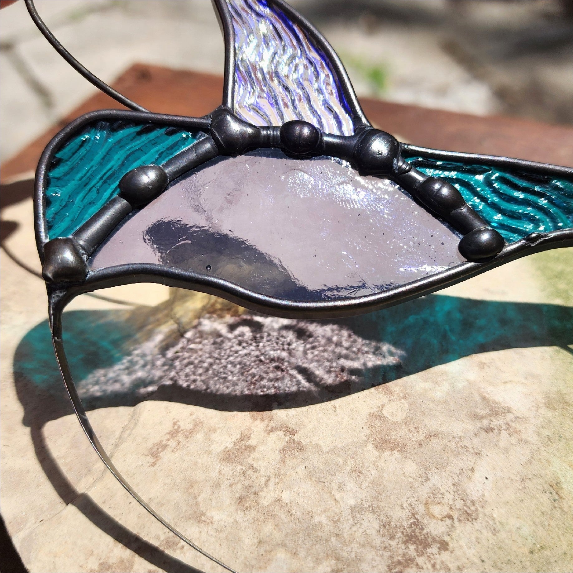 A stained glass hummingbird suncatcher featuring textured teal and purple-blue iridescent wing, teal head and tail, a central lavender body, and dark metal lead lines, resting on a stone surface outdoors.