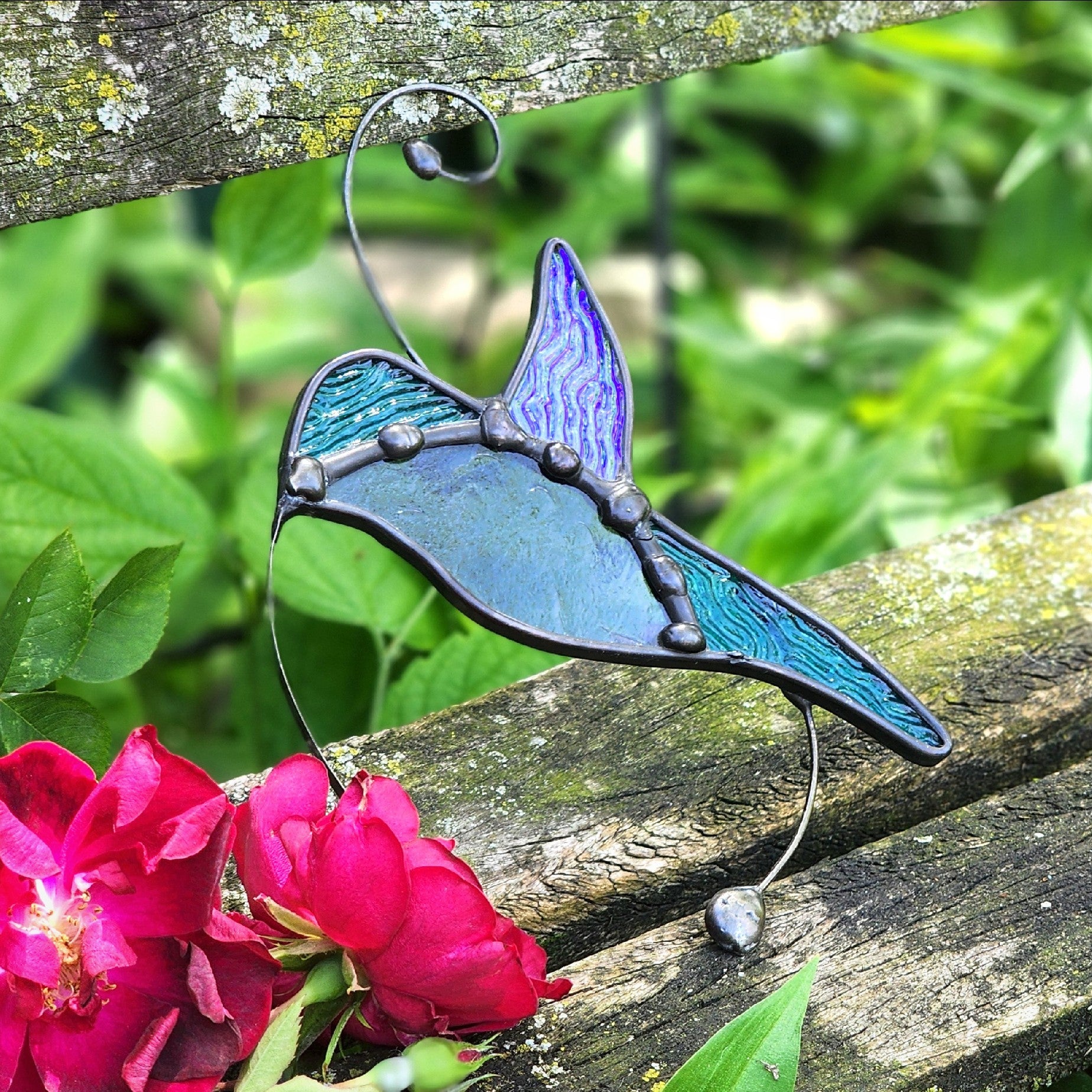 A stained glass hummingbird suncatcher with teal and purple iridescent glass panels, held in place by a metal wire frame, resting on a weathered wooden bench with green foliage and pink flowers in the background.