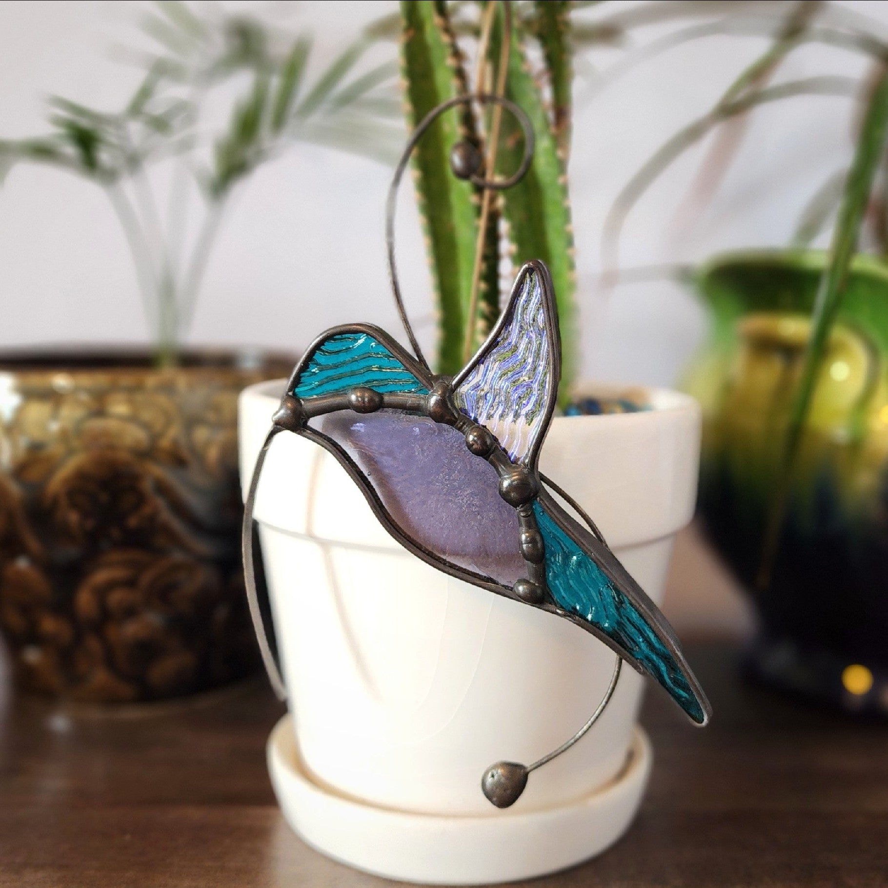 A stained glass hummingbird suncatcher with teal, purple, and iridescent blue glass glass, suspended in a white ceramic planter on a wooden table with potted plants in the background.
